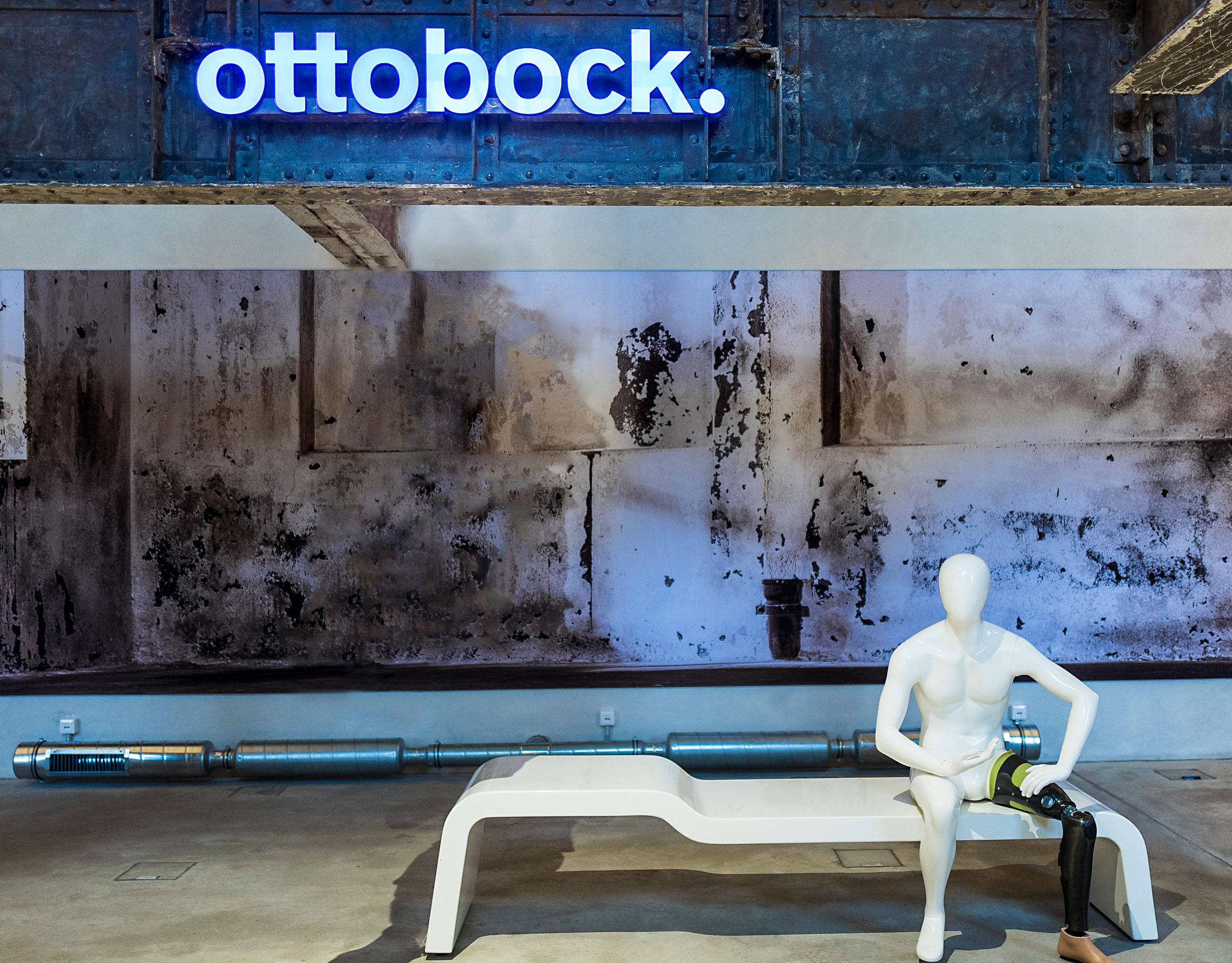 Weiße Skulptur mit Beinprothese auf einer Bank vor bläulichem Hintergrund mit Leuchtschrift "Ottobock"